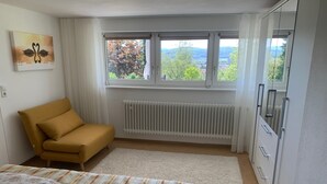 A bedroom with a sofa bed, a radiator, a window with curtains, and a view of trees and hills.