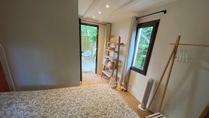 A bedroom with a patterned bedspread, a wooden ladder, a shelf with towels, and a view through a window.