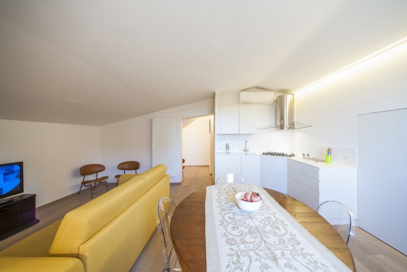A modern living room with a yellow sofa, a dining table with a white tablecloth, and a kitchen area with white cabinets and a stainless steel range hood.
