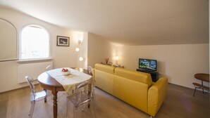 Een eetgedeelte met een ronde tafel, stoelen, een gele bank, een televisie en een raam met natuurlijk licht.