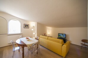 Ein Essbereich mit einem runden Tisch, Stühlen, einem gelben Sofa, einem Fernseher und einem Fenster mit Tageslicht.