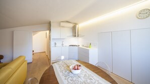 Een kleine keuken met witte kastjes, een eettafel met een tafelkleed met dessin, en een schaal fruit.