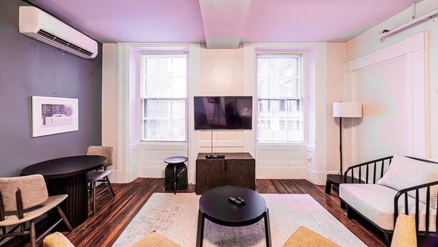 Uma sala de estar moderna com uma televisão de tela plana, duas janelas com persianas, um sofá, uma mesa de centro e uma pequena mesa lateral.