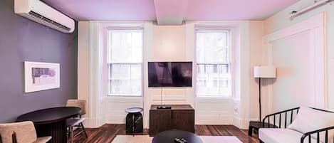 Una sala de estar moderna con un televisor de pantalla plana, dos ventanas con persianas, un sofá, una mesa de centro y una mesita auxiliar.