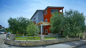 A modern two-story building with a red and grey facade, a covered outdoor seating area, and a landscaped garden with trees and a brick pathway. - Coastal Serene Stay in Umag (Umag)