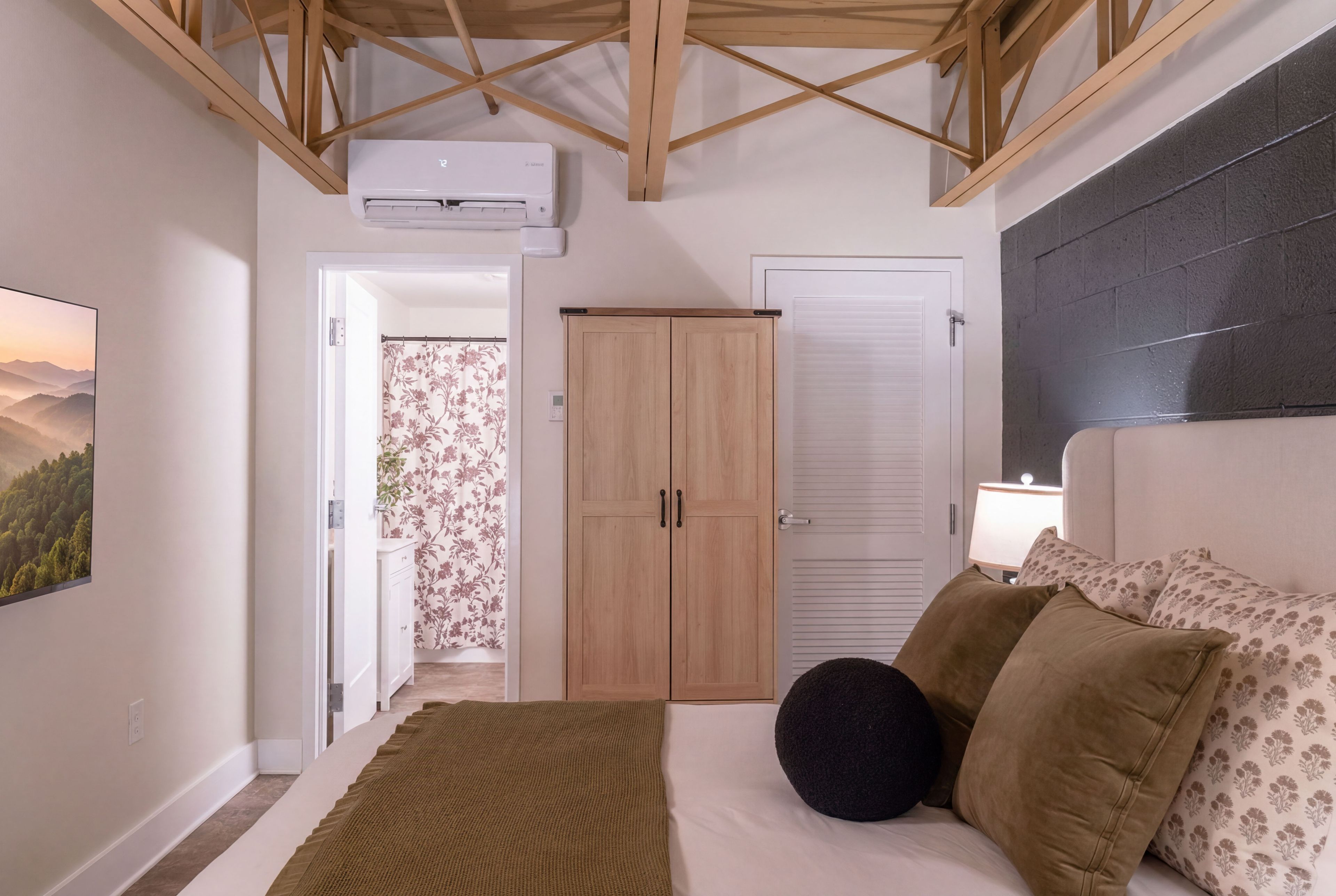 A bedroom with a bed, pillows, a nightstand, a wall-mounted lamp, a wardrobe, and an air conditioning unit.
