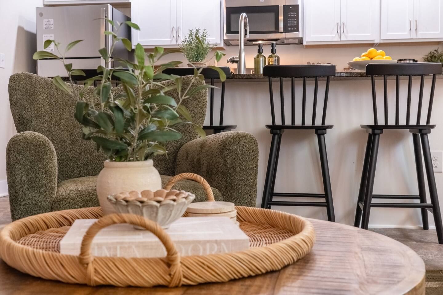 Une cuisine moderne avec des meubles blancs, un réfrigérateur en acier inoxydable, un four à micro-ondes et un évier. On y trouve deux tabourets de bar noirs et une table ronde en bois sur laquelle reposent un plateau en osier et un vase avec des plantes vertes.