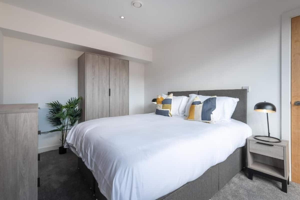 A modern bedroom with a bed, bedside table, lamp, wardrobe, and a potted plant.