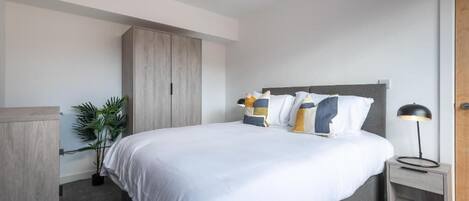 A modern bedroom with a bed, bedside table, lamp, wardrobe, and a potted plant.