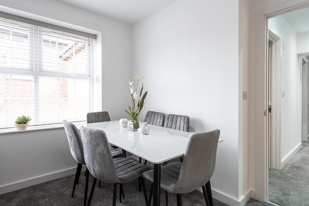 A dining area with a white table and grey upholstered chairs, a potted plant, and a window with blinds.