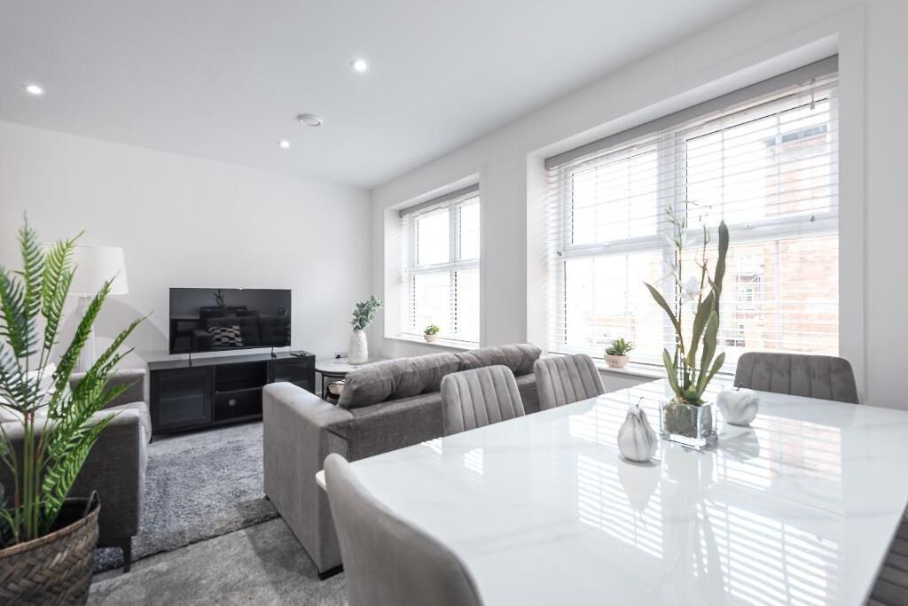 Una sala de estar moderna con un televisor de pantalla plana, sofá gris, mesa de comedor y ventanas amplias.