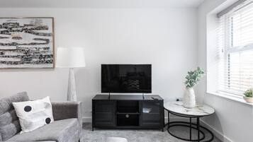 Ein Wohnzimmer mit einer Couch, einem Fernseher, einer Lampe, einem kleinen Tisch und einem Fenster mit Jalousien.