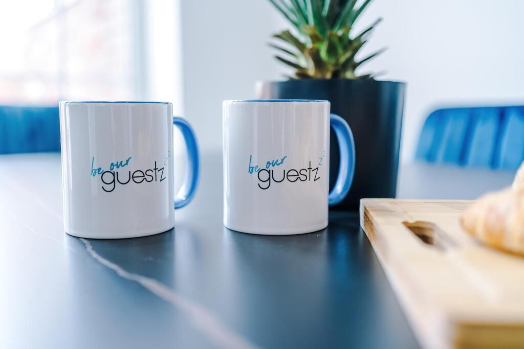 Two white mugs with 'be our guest' text on a table.