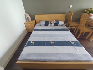 Cama bem arrumada com colcha listrada, cabeceira de madeira, mesa de cabeceira com luminária e porta-retrato, e uma cadeira de madeira.