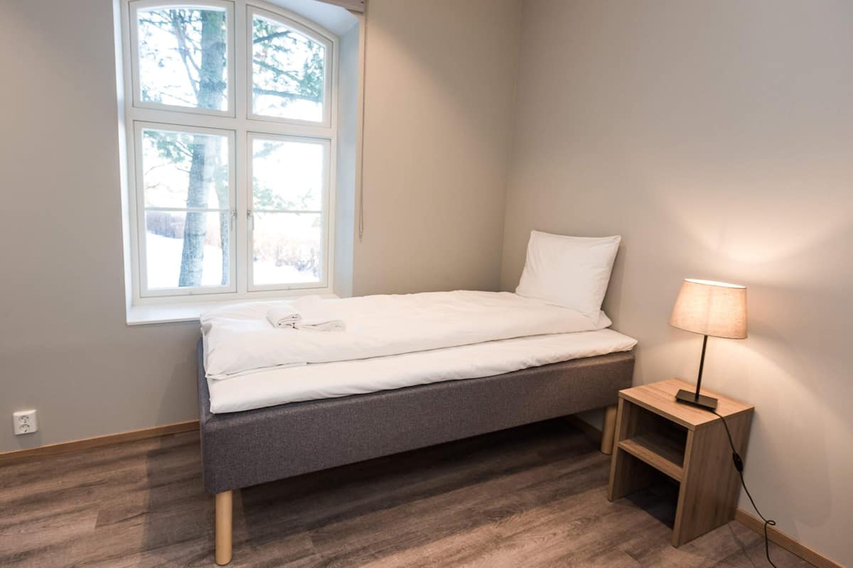 Uma cama de solteiro com estrutura cinzenta, roupa de cama branca, uma pequena mesa de cabeceira com abajur e uma janela com vista para árvores.