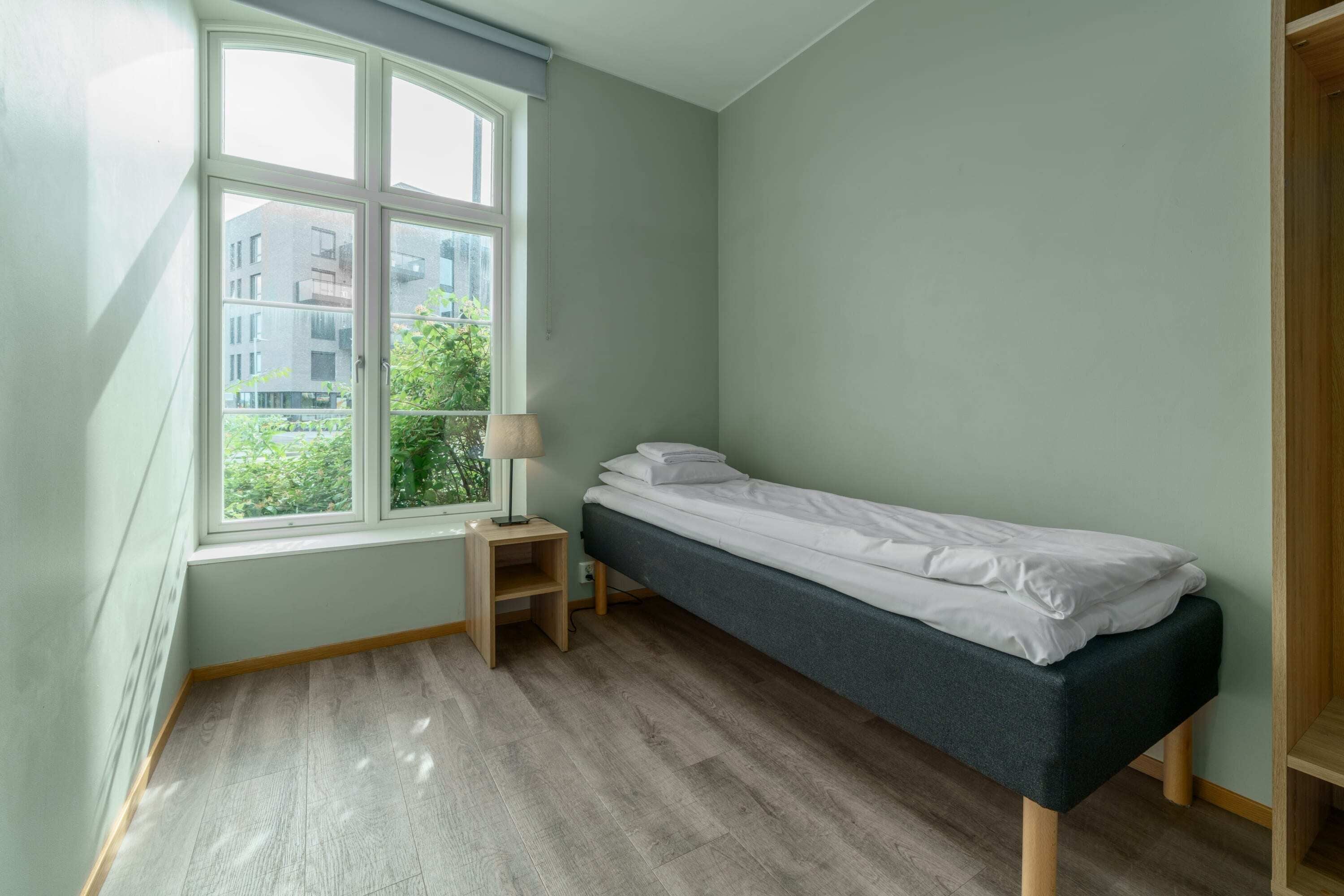 A small, single-bed room with a window, a nightstand, and a wardrobe.