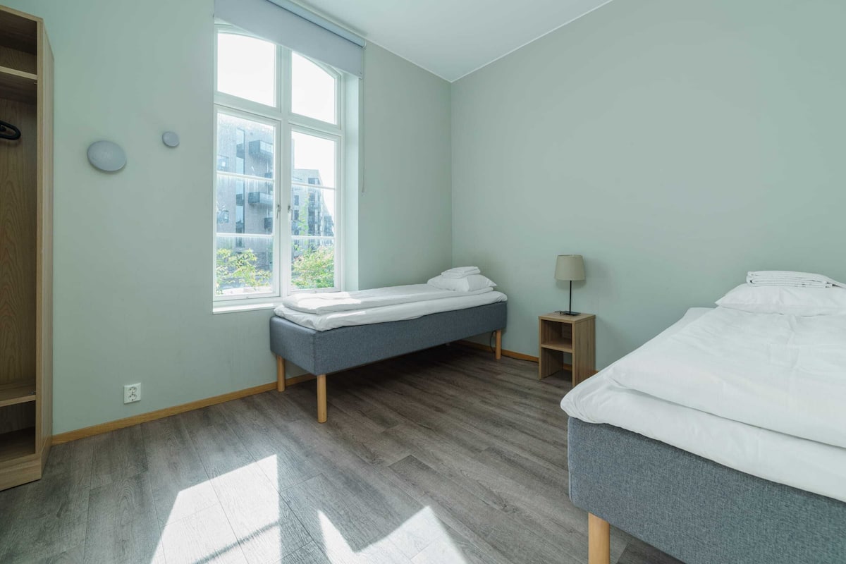 Um quarto com uma cama, mesa de cabeceira, abajur e janela com vista para prédios.