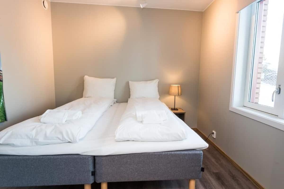 Uma cama de casal com lençóis brancos, uma luminária de cabeceira e uma janela com cortinas.