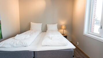 Una cama doble con sábanas blancas, una lámpara de noche y una ventana con persianas.