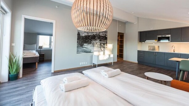 Una habitación de hotel moderna con una cama grande, un escritorio pequeño, una kitchenette y un ventanal amplio.