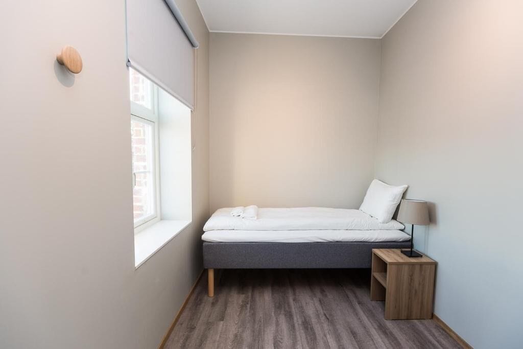 Una habitación pequeña con cama individual, ventana, mesita de noche con lámpara y suelo de madera.