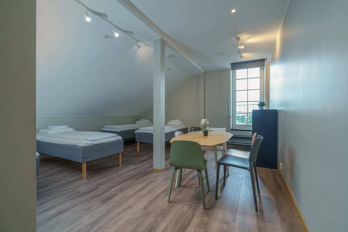 Quarto estilo dormitório com beliches, uma pequena mesa, cadeiras e uma janela.