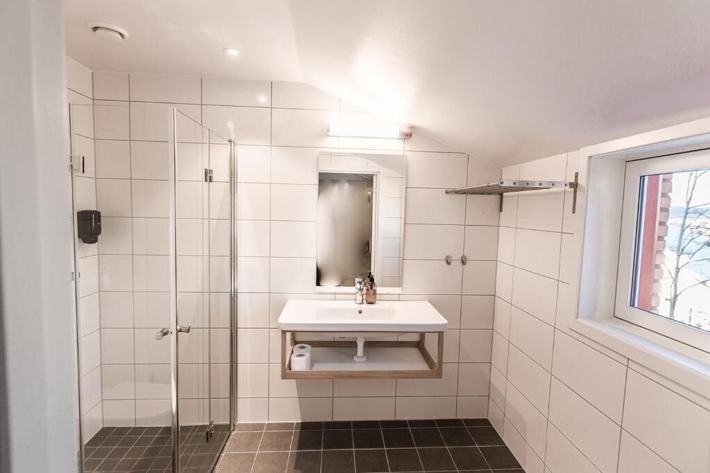 A bathroom with a glass shower enclosure, a white sink with a mirror above, and a window to the right.