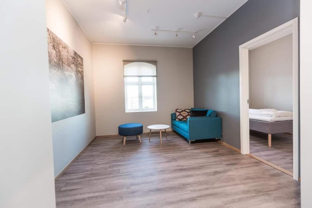 Quarto com chão de madeira, um sofá azul, uma mesa redonda pequena, uma janela e uma cama.