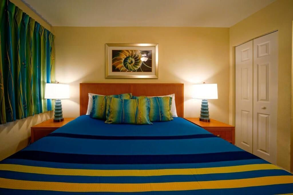 Ein Hotelzimmer mit einem Bett, zwei Nachttischlampen, einem gerahmten Kunstwerk über dem Bett und einer Tür rechts.