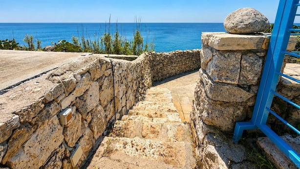 Ein steiniger Weg führt zu einer Steinmauer mit Blick auf das Meer.