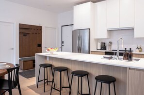 A modern kitchen with a central island, bar stools, a refrigerator, and white cabinetry. - Bright & stylish 2-bedroom, 2-bath condo in Mackenzie Village—your cozy mountain retreat, just minutes from outdoor adventure, dining, and year-round recreation. (Revelstoke)