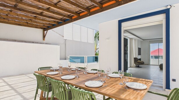 Un área de comedor al aire libre con una mesa de madera preparada para seis personas, rodeada de sillas verdes, bajo una pérgola de madera.