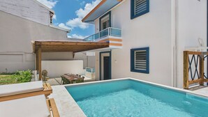 Una piscina con sillones de descanso, una pérgola de madera y un edificio de dos plantas con balcones.