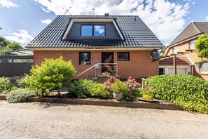 Une maison à deux étages avec une façade en briques, un toit en tuiles et un jardin devant, agrémenté de diverses plantes et fleurs.