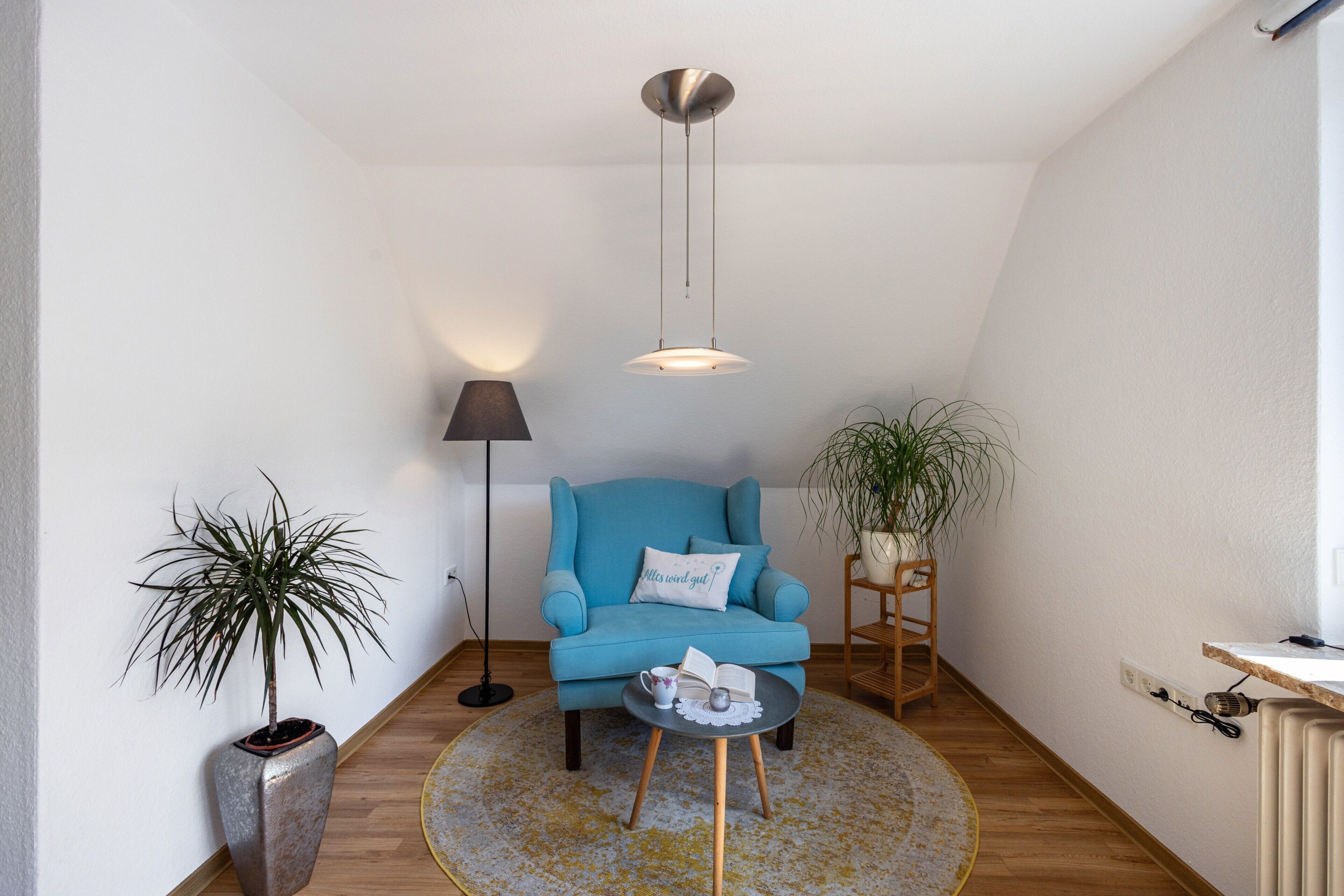 Une pièce chaleureuse avec un fauteuil bleu, une petite table sur laquelle se trouvent un livre et une tasse, un lampadaire et deux plantes en pot.