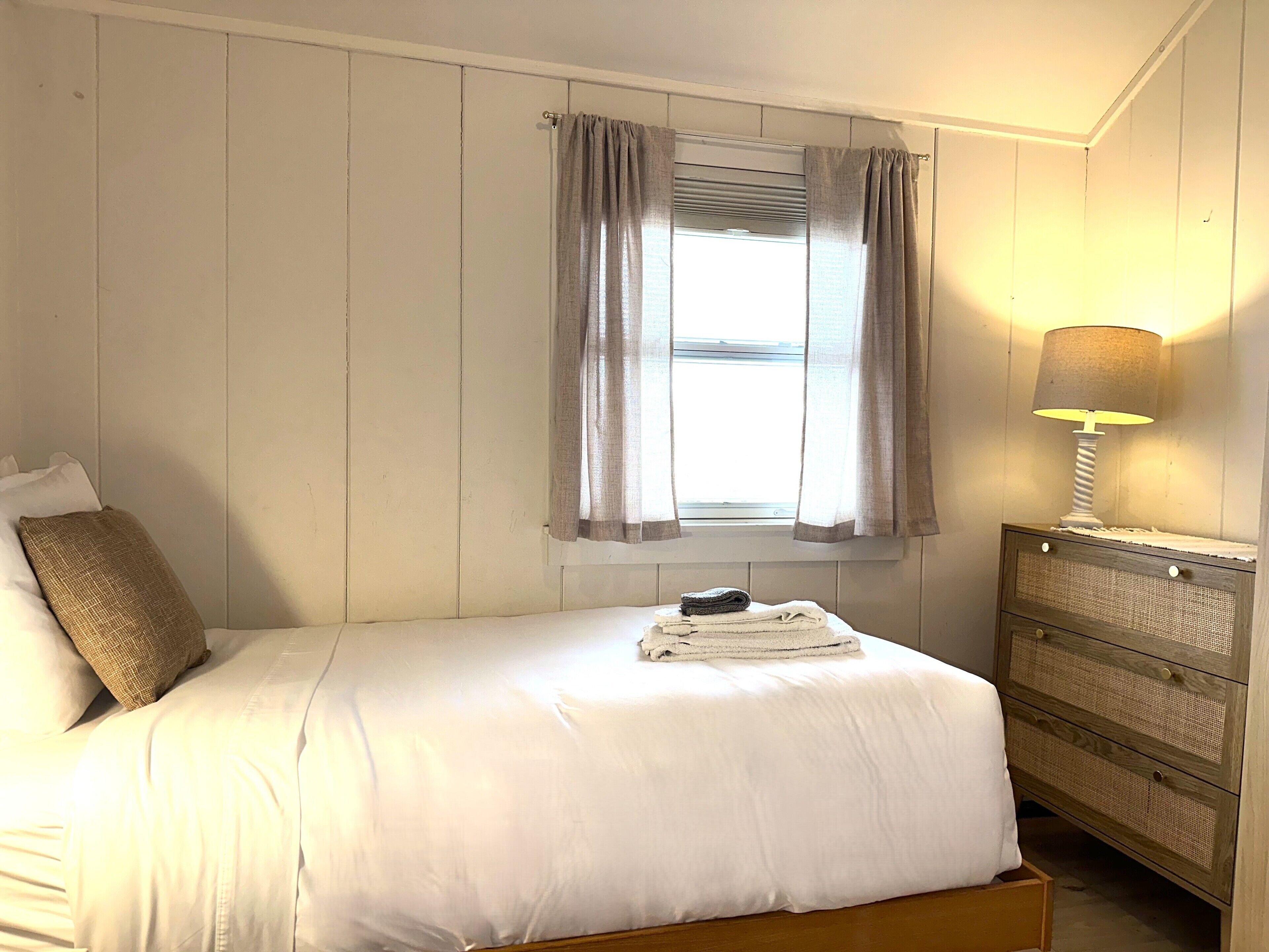 A bedroom with a bed, a window with curtains, a bedside lamp, and a wooden dresser.