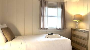 A bedroom with a bed, a window with curtains, a bedside lamp, and a wooden dresser.