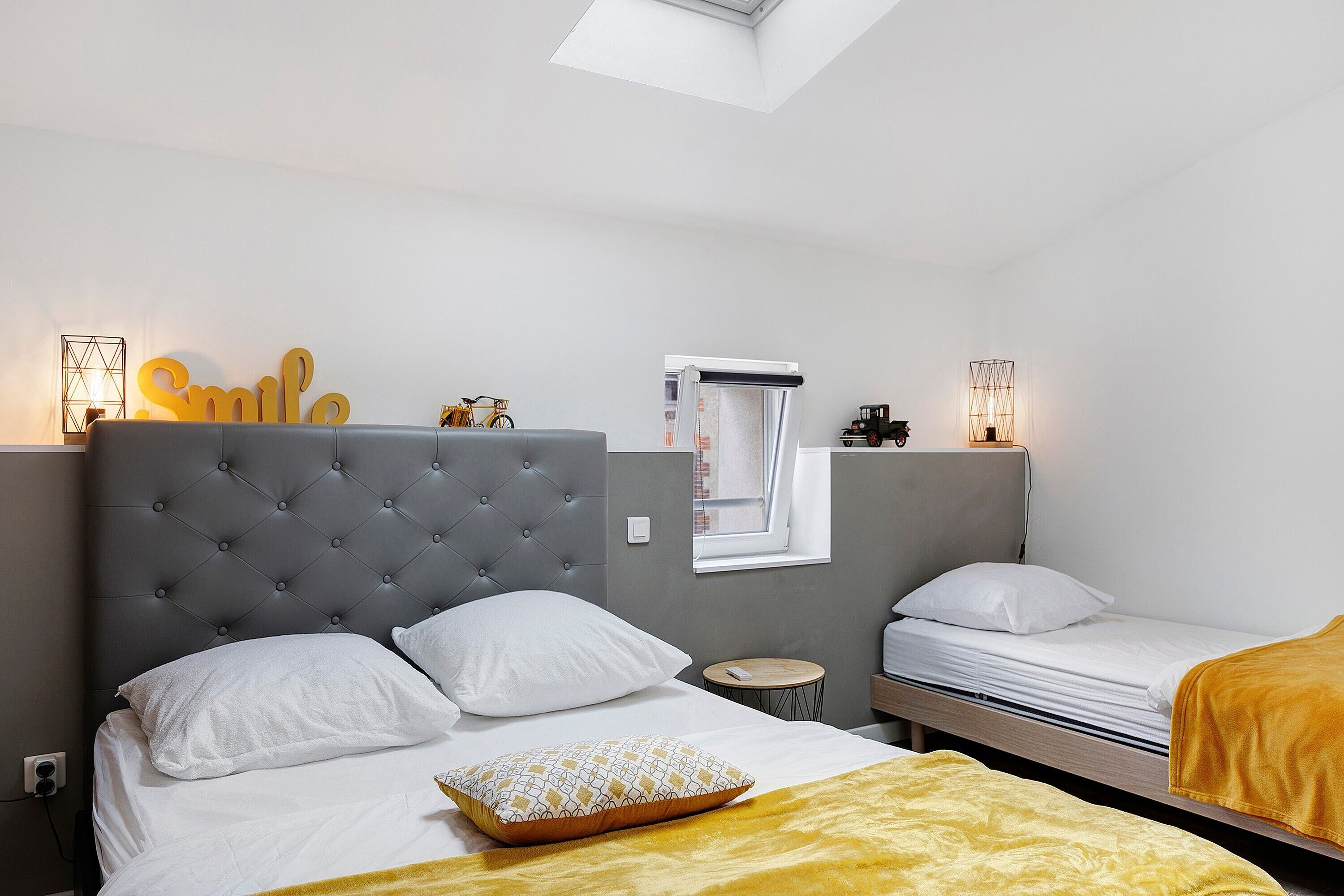Habitación de hotel con tablero de cabecera capitonado, cama con ropa de cama blanca, una pequeña mesa redonda, una ventana con vistas y un cartel con la palabra 'Smile' en la pared.