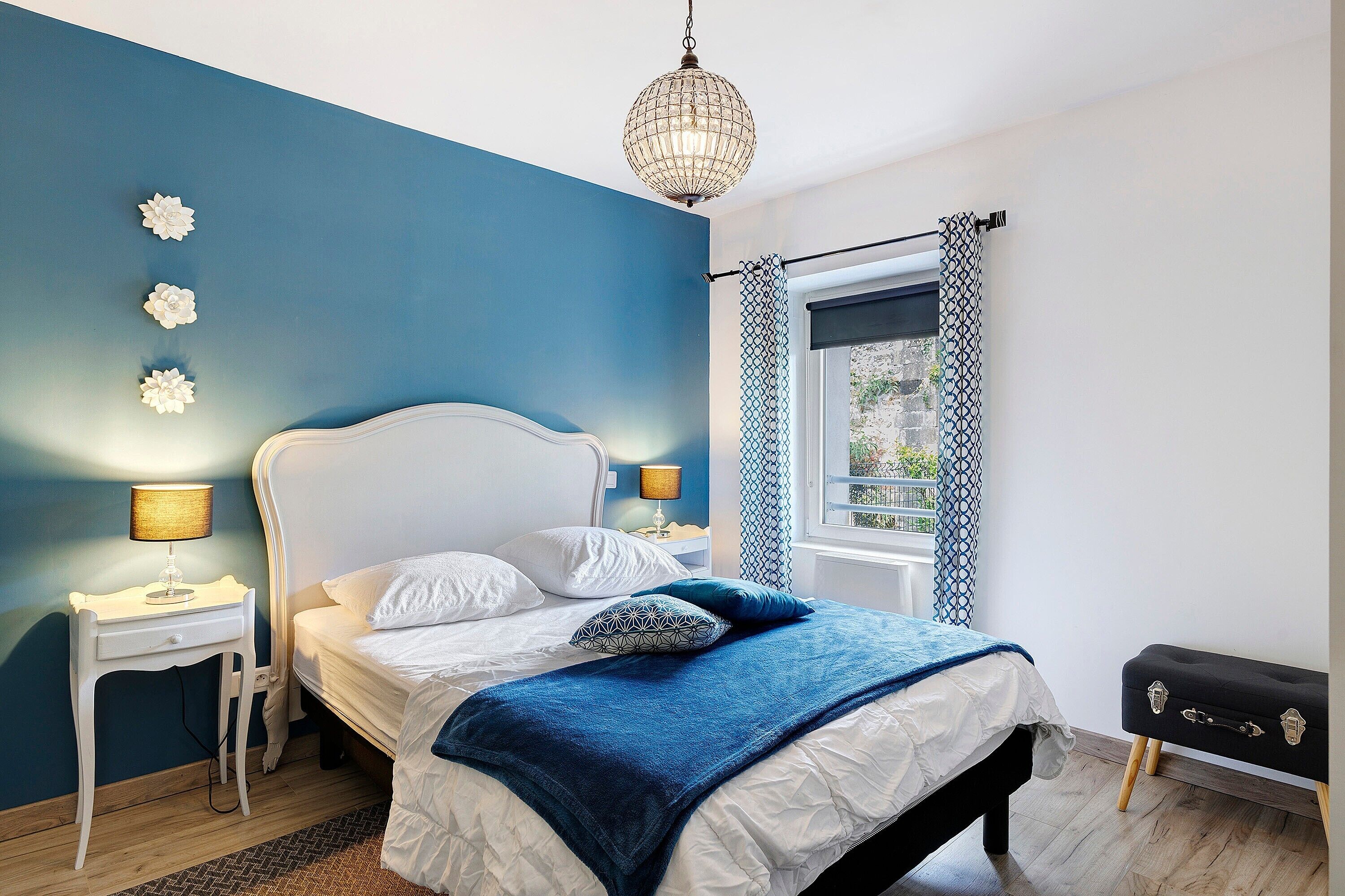 A bedroom with a bed, bedside tables, a lamp, a decorative wall piece, a window with curtains, and a suitcase.