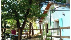 A wooden deck with a table and chairs, a small building with a blue door, and a tree in the background. - Small house in the heart of the Cévennes (Concoules)