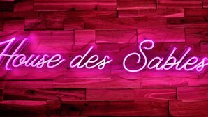 Een neonbord met de tekst 'House des Sables' op een houten wand.