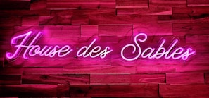 Ein Neon-Schild mit dem Schriftzug 'House des Sables' an einer Holzverkleidung.
