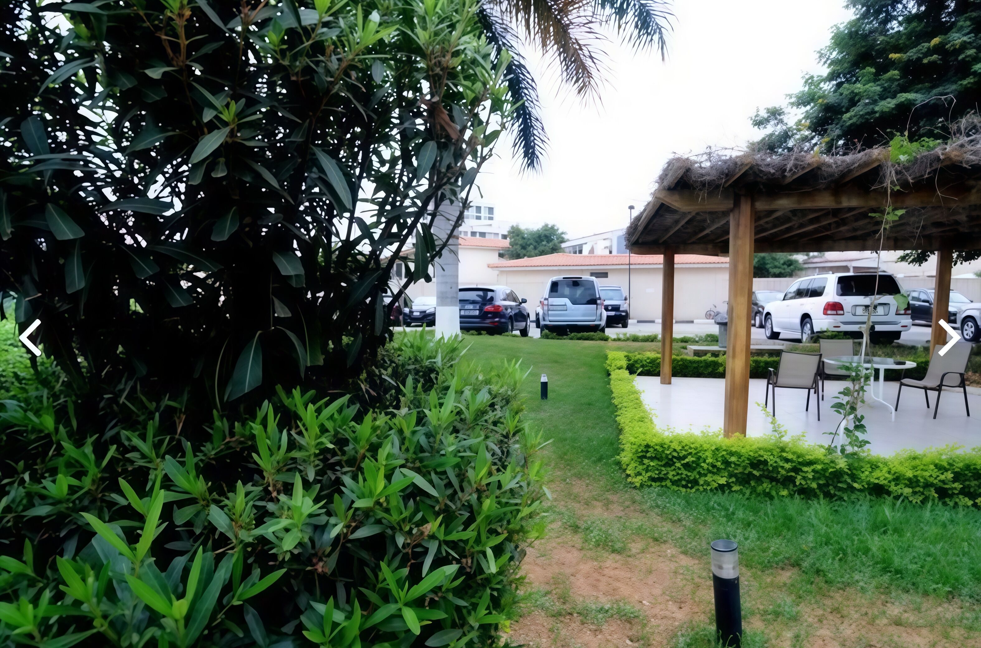 A pathway leading to a covered outdoor seating area with a thatched roof, surrounded by greenery and parked cars.