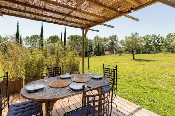 Mesa de madera al aire libre dispuesta para comer, con sillas, una pérgola arriba y un campo de hierba al fondo.