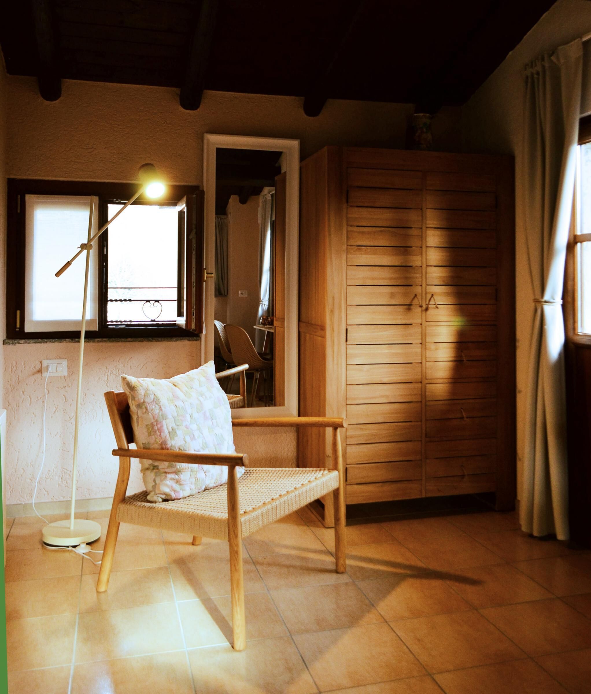 Una stanza con una sedia di legno, una lampada da terra, un armadio e una finestra con tende.