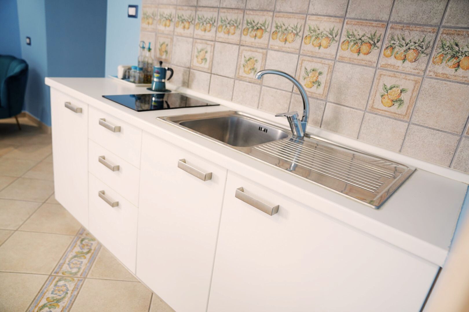A kitchen with a stainless steel sink, white cabinets, and a tiled backsplash with fruit designs.