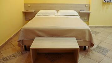 Una cama con cabecera de madera, una mesita de noche y un banco en una habitación con paredes amarillas y suelo de baldosas estampadas.