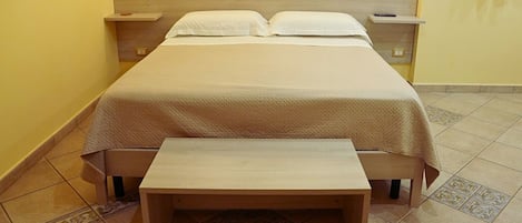 A bed with a wooden headboard, a bedside table, and a bench in a room with yellow walls and patterned floor tiles.