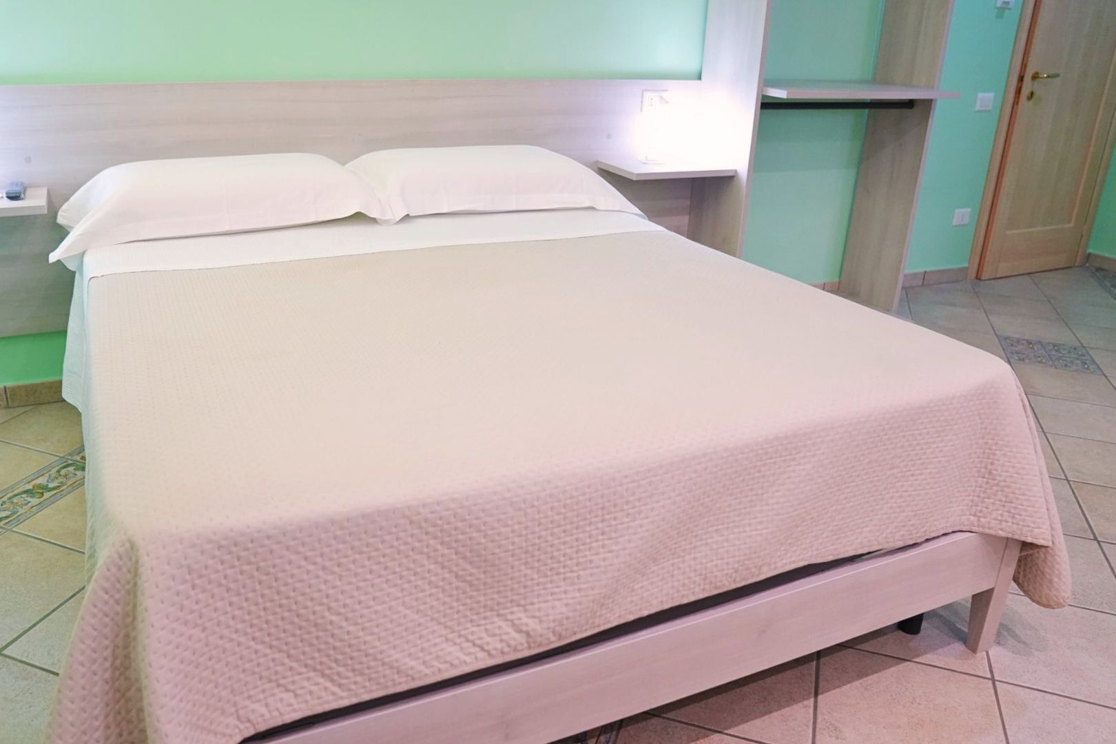 Un lit avec un matelas blanc et une couverture à motifs, une table de chevet et une étagère fixée au mur.