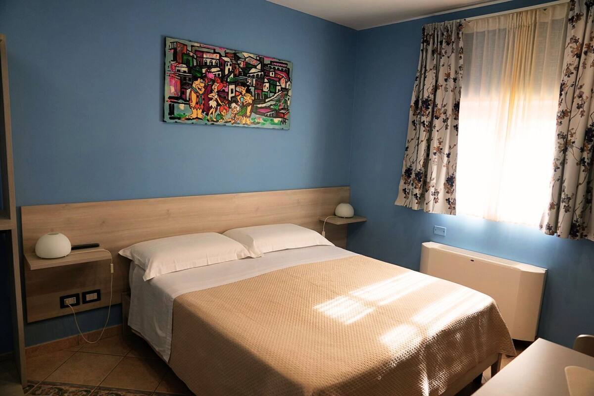 A bedroom with a bed, bedside tables, a wall-mounted lamp, a radiator, and a window with curtains.